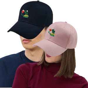 Puede incluir: Dos gorras de béisbol, una azul marino y otra rosa claro, cada una bordada con un diseño de Kermit la Rana. Las gorras tienen una visera curva y una correa ajustable. Un accesorio divertido.