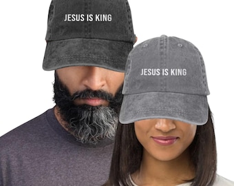 May include: Two gray denim baseball caps with the text "JESUS IS KING" in white. The caps are worn by two people. The caps have a curved brim and an adjustable strap at the back.
