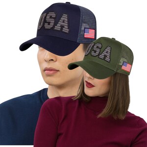 May include: Two baseball caps with the word "USA" in a block letter design. One cap is navy blue with a gray mesh back and a small American flag patch. The other is olive green with a gray mesh back and a small American flag patch.