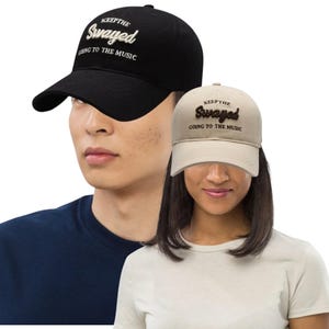 May include: Two baseball caps, one black and one tan, with the text "KEEP THE SWAYED GOING TO THE MUSIC" embroidered on the front. The black cap is worn by a person wearing a navy blue shirt. The tan cap is worn by a person wearing a white shirt.