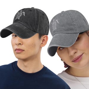 May include: Two baseball caps are shown, one dark grey and one light grey. Each cap has a white embroidered sea turtle design on the front. The caps have a classic, adjustable design, suitable for various head sizes.