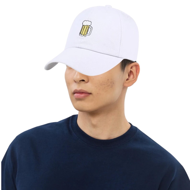 May include: White baseball cap with a curved brim. The cap features an embroidered design of a beer mug with yellow and brown details. The cap is worn by a person wearing a navy blue shirt.