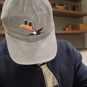 May include: Gray baseball cap featuring a toucan graphic with a black body, orange beak, and yellow and black accents. The toucan is depicted in flight. The cap is made of a soft material and has a curved brim.