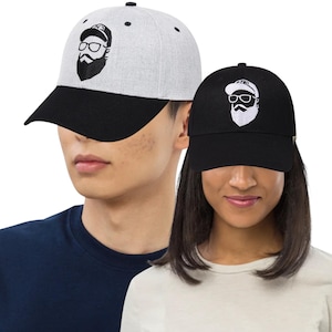 May include: Two baseball caps, one light gray and one black, each with a black brim. Both caps feature an embroidered design of a bearded face wearing glasses. The caps are worn by two people.