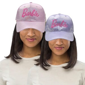 Könnte beinhalten: Zwei Baseballkappen, eine hellrosa und eine lavendelfarbene, jeweils mit dem Namen "Barbie" in einer kontrastierenden Farbe bestickt. Die Kappen haben ein glänzendes, glitzerndes Finish und einen gebogenen Schirm.