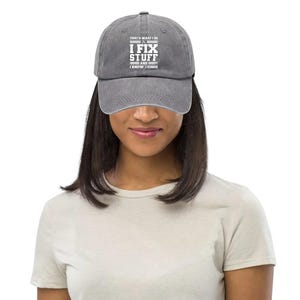 Könnte beinhalten: Graue Baseballkappe mit dem weißen Text: "THAT'S WHAT I DO I FIX STUFF AND I KNOW THINGS". Die Kappe hat einen gebogenen Schirm und einen verstellbaren Riemen. Der Hut ist aus einem weichen, abgenutzten Material.