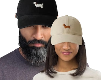 Puede incluir: Dos gorras de béisbol, una negra y otra beige, cada una bordada con un perro salchicha. La gorra negra tiene un perro blanco y marrón, mientras que la gorra beige presenta un perro marrón. Ambas gorras son usadas por personas.