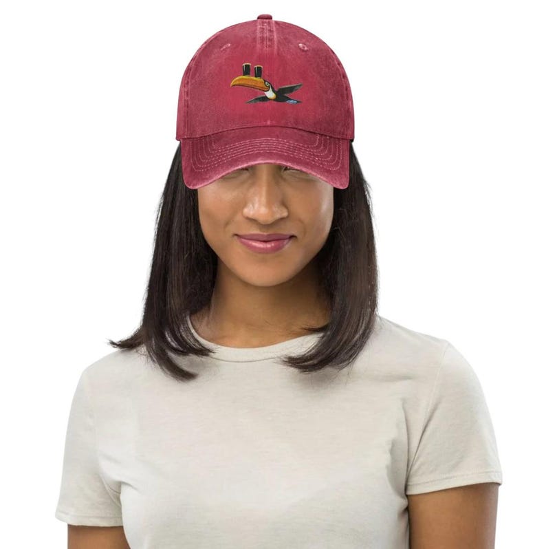 K&ouml;nnte beinhalten: Burgunderfarbene Baseballkappe mit einem Tukan-Motiv. Der Tukan hat einen gelben Schnabel und wird im Flug dargestellt. Die Kappe besteht aus einem weichen Material und hat einen gebogenen Schirm. Vintage-Stil.