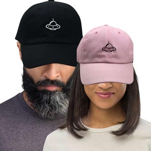 Puede incluir: Dos gorras de béisbol, una negra y otra rosa, cada una bordada con un diseño de OVNI blanco. La gorra negra la lleva una persona con barba, y la gorra rosa la lleva una persona con pelo largo y oscuro. Las gorras son de estilo casual.