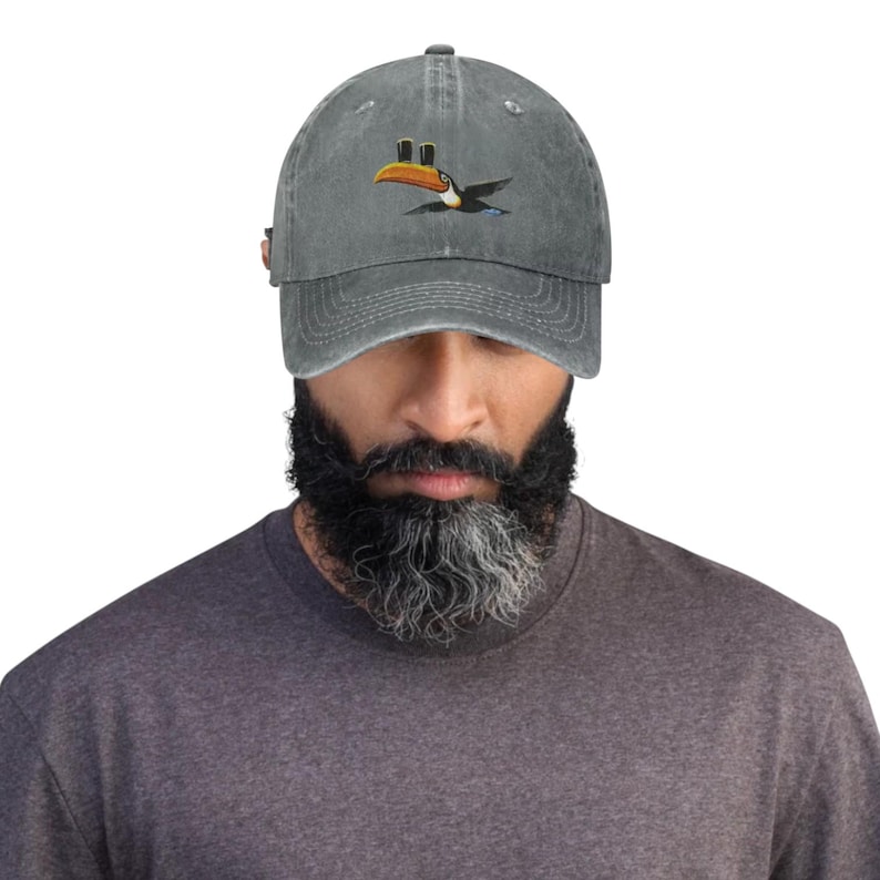 K&ouml;nnte beinhalten: Graue Baseballkappe mit Used-Look. Die Kappe zeigt einen gestickten Tukan mit gelbem Schnabel und schwarzen und blauen Fl&uuml;geln. Der Tukan fliegt. Die Kappe hat einen gebogenen Schirm und einen verstellbaren Riemen hinten.