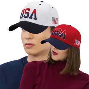 May include: Two baseball caps with the word "USA" embroidered in red and navy blue. One cap is white with a black brim, and the other is red with a navy blue brim. Both caps feature a small American flag on the side.