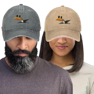 K&ouml;nnte beinhalten: Zwei Baseballkappen, eine graue und eine beige, jeweils mit einem gestickten Tukan-Design. Der Tukan hat einen gro&szlig;en orangefarbenen Schnabel und schwarze Fl&uuml;gel. Die Kappen sind aus weichem, abgenutztem Material.