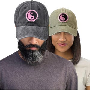 Puede incluir: Dos gorras de béisbol, una gris antracita y otra verde oliva, cada una con un parche de corazón yin-yang rosa y negro. Las gorras las llevan dos personas. El fondo es blanco. Un accesorio casual.