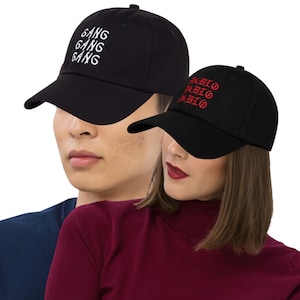 Könnte beinhalten: Zwei schwarze Baseballkappen. Auf einer Kappe ist das Wort "GANG" dreimal weiß gestickt. Auf der anderen Kappe ist das Wort "FABIO" dreimal rot gestickt. Beide Kappen haben einen gebogenen Schirm und einen verstellbaren Riemen.