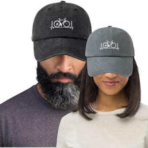 Puede incluir: Dos gorras de béisbol, una negra y otra gris, cada una con un gráfico de bicicleta blanco y una línea de latido cardíaco. Las gorras están hechas de un material suave y texturizado y tienen una visera curva. Llevadas por dos personas.