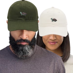 Pode incluir: Dois bonés de beisebol, um verde-oliva e outro branco-sujo, cada um bordado com um pequeno desenho de tartaruga. Os bonés são feitos de um tecido macio e têm uma aba curva. Um acessório casual.
