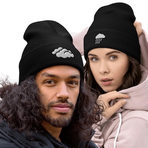 May include: Two black beanies with white cloud embroidery. One beanie features a cloud with rain, the other has a cloud design. The beanies are worn by two people. The beanies are made of a soft, knitted material.