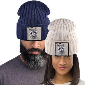 Könnte beinhalten: Zwei gerippte Strickmützen, eine marineblaue und eine beige, jeweils mit einem quadratischen Aufnäher mit einer Waschbär-Illustration und dem Wort "RACCOON". Die Mützen werden von zwei Personen getragen.