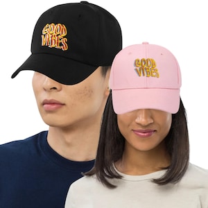 May include: Two baseball caps, one black and one pink, each with the embroidered text "GOOD VIBES" in a colorful, retro font. The caps have a curved brim and a classic six-panel design. The black cap is worn by a person wearing a navy blue shirt, and the pink cap is worn by a person wearing a white shirt.