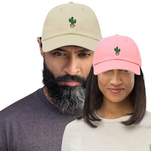 Könnte beinhalten: Zwei Baseballkappen, eine beige und eine rosa, jeweils mit einem grünen Kaktus-Design auf der Vorderseite gestickt. Die Kappen werden von zwei Personen getragen. Die beige Kappe wird von einem Mann mit dunklem Bart getragen, und die rosa Kappe wird von einer Frau getragen.