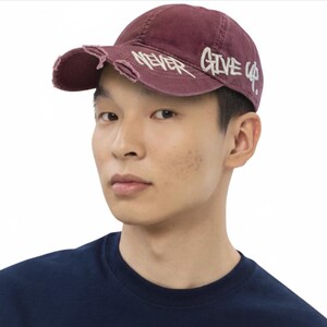 Könnte beinhalten: Burgunderfarbene Baseballkappe mit Used-Details und dem weißen Schriftzug "NEVER GIVE UP". Die Kappe hat einen gebogenen Schirm und ist aus weichem, abgenutztem Material.
