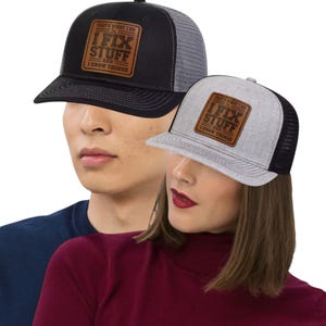 Könnte beinhalten: Zwei Trucker-Hüte, einer schwarz und einer grau, mit Mesh-Rücken und gebogenen Krempen. Jeder Hut hat einen braunen Lederaufnäher mit dem Text "THAT'S WHAT I DO, I FIX STUFF AND I KNOW THINGS". Die Hüte werden von zwei Personen getragen.