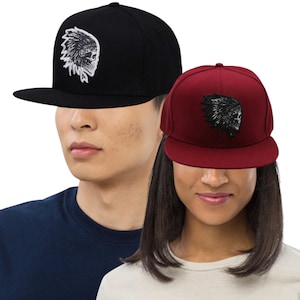 Op de afbeelding: Twee baseballpetten met platte klep. Eén is zwart met een wit en zwart geborduurd ontwerp van een Indiaanse hoofdtooi en schedel. De andere is bordeauxrood met een zwart geborduurd ontwerp van een Indiaanse hoofdtooi en schedel.