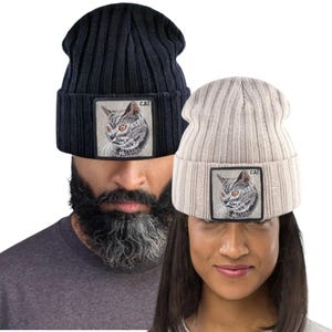 Könnte beinhalten: Zwei gerippte Strickmützen. Eine ist marineblau, die andere beige. Jede Mütze hat einen quadratischen Aufnäher mit einer detaillierten Illustration einer Katze mit orangefarbenen Augen und dem Wort "CAT".