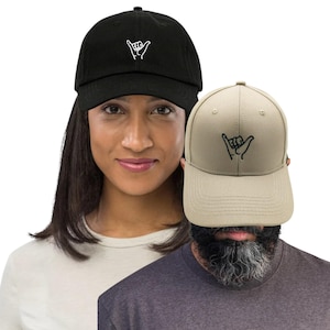 Può includere: Due cappellini da baseball, uno nero e uno beige, ciascuno con un gesto della mano ricamato in bianco. Il cappellino nero è indossato da una donna e quello beige da un uomo. Entrambi i cappellini hanno una visiera curva e un cinturino regolabile.