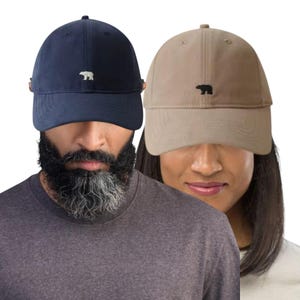 May include: Two baseball caps, one navy blue and one beige, each with a small bear logo embroidered on the front. The caps are worn by two people, partially obscuring their faces. The caps are a casual accessory.