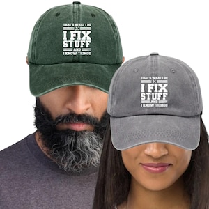 Könnte beinhalten: Zwei Baseballkappen, eine dunkelgrüne und eine graue, mit dem Text "THAT'S WHAT I DO, I FIX STUFF, AND I KNOW THINGS" in Weiß. Die Kappen werden von zwei Personen getragen.