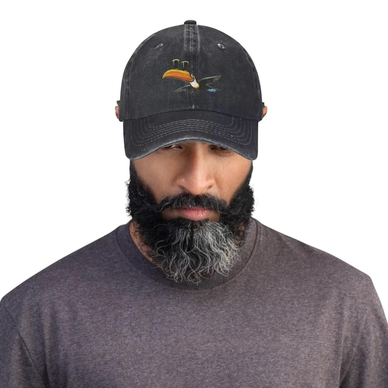K&ouml;nnte beinhalten: Dunkelgraue Baseballkappe mit Used-Look. Auf der Kappe ist ein gesticktes Design eines Tukanvogels mit orangefarbenen, gelben und blauen Akzenten zu sehen. Die Kappe hat einen gebogenen Schirm und einen verstellbaren Riemen.