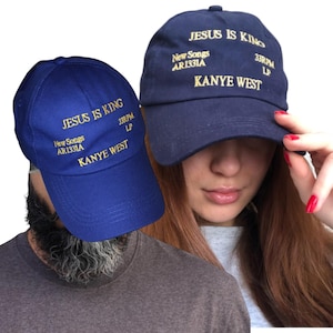 Könnte beinhalten: Zwei marineblaue Baseballkappen mit goldfarbenem, gesticktem Text. Der Text auf den Kappen lautet "JESUS IS KING", "New Songs AR1331A", "33RPM LP" und "KANYE WEST". Die Kappen werden vor weißem Hintergrund gezeigt.
