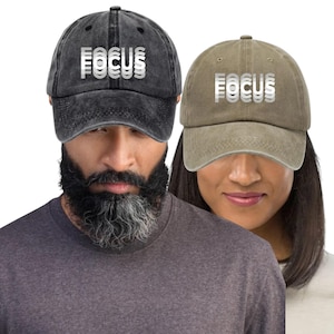 Peut inclure: Deux casquettes de baseball avec le mot "FOCUS" en blanc, texte superposé. Une casquette est noire, l'autre est vert olive. Les casquettes sont portées par deux personnes, l'une avec une barbe et l'autre avec de longs cheveux.