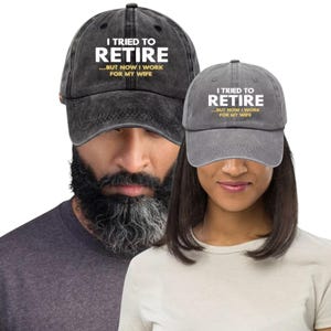 May include: Two gray baseball caps with the text "I TRIED TO RETIRE...BUT NOW I WORK FOR MY WIFE" in white and gold. The caps are worn by a person with a beard and a person with short hair.
