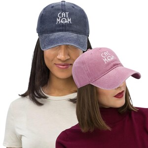 May include: Two baseball caps, one navy blue and one dusty rose, are displayed. Both caps have the embroidered text "CAT MOM" in white. The caps are made of a soft, textured fabric and have a curved brim.