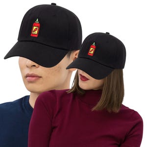 May include: Black baseball cap with a curved brim. The cap features an embroidered design of a red hot sauce bottle with a green top and the word "HOT" inside. The cap is worn by two people, one wearing a navy blue shirt and the other wearing a burgundy turtleneck.
