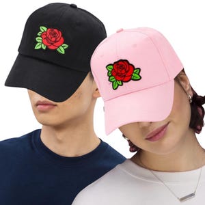 May include: Two baseball caps, one black and one pink, each featuring an embroidered red rose with green leaves. The caps have a curved brim and a classic six-panel design, suitable for casual wear.
