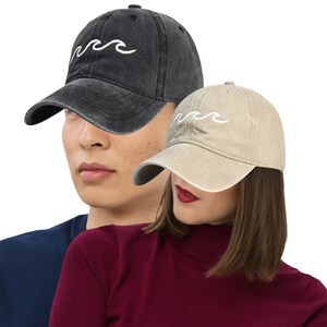 Könnte beinhalten: Zwei Baseballkappen, eine schwarze und eine beige, jeweils mit einem weißen, gestickten Wellenmuster. Die Kappen werden von zwei Personen getragen, eine mit einem marineblauen Oberteil und die andere mit einem bordeauxfarbenen Rollkragenpullover.