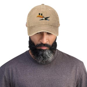 K&ouml;nnte beinhalten: Beige Baseball Cap mit gebogenem Schirm. Die Kappe hat ein gesticktes Tukan-Vogel-Design in Gelb, Orange, Schwarz und Blau. Die Kappe ist aus einem weichen, abgenutzten Material gefertigt und hat einen verstellbaren Riemen.