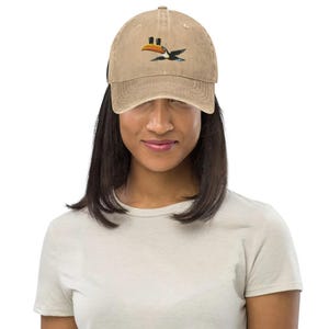 K&ouml;nnte beinhalten: Beige Baseball Cap mit gebogenem Schirm. Die Kappe hat ein gesticktes Tukan-Design in Orange, Schwarz und Blau. Die Kappe wird von einer Person getragen, die ein helles T-Shirt tr&auml;gt.