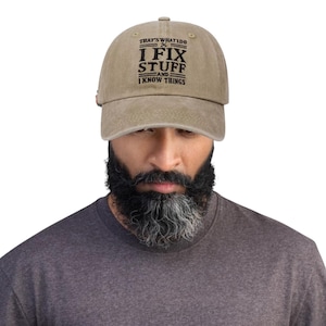May include: A tan baseball cap with the text "THAT'S WHAT I DO I FIX STUFF AND I KNOW THINGS" in black lettering. The cap has a curved brim and a fabric strap closure. The hat is worn by a person.