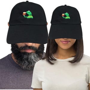 May include: Two black baseball caps with a Kermit the Frog embroidery design. The embroidered Kermit is green and holding a brown beverage. The caps are worn by two people.