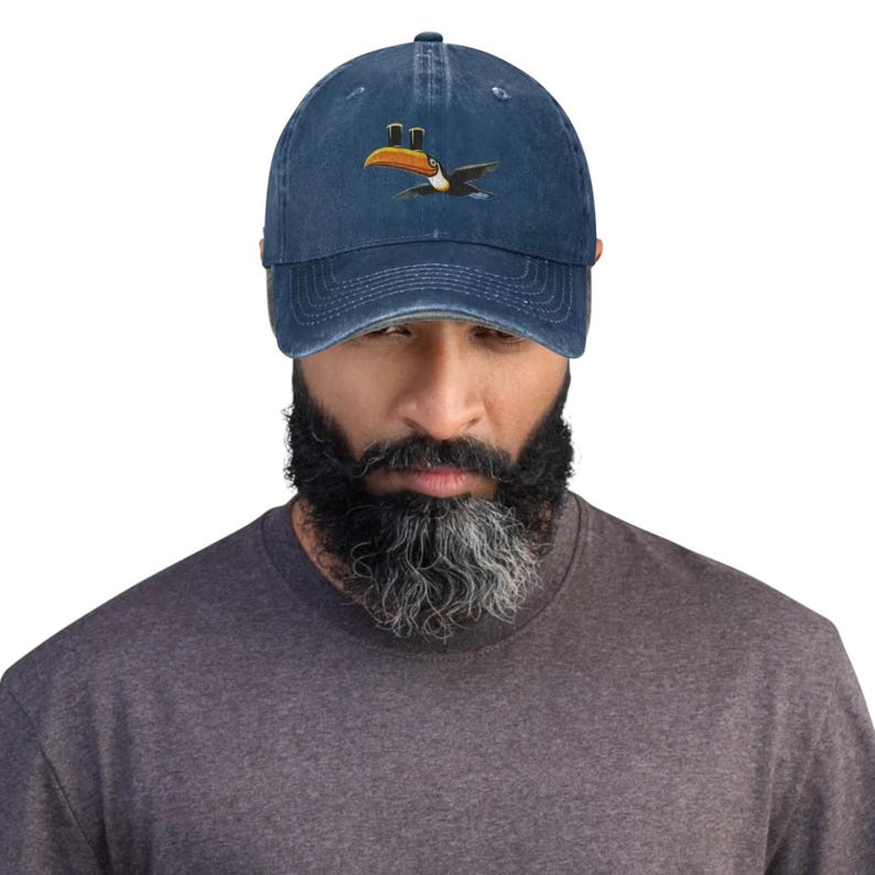 K&ouml;nnte beinhalten: Marineblaue Baseballkappe mit Used-Look. Die Kappe zeigt einen gestickten Tukan mit Zylinder. Der Tukan hat einen gro&szlig;en orangefarbenen Schnabel, schwarze Fl&uuml;gel und einen wei&szlig;en Bauch. Die Kappe hat einen gebogenen Schirm und einen verstellbaren Riemen.