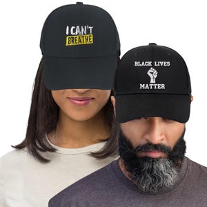 Może przedstawiać: Dwie czarne czapki z daszkiem. Jedna czapka ma napis "I CAN'T BREATHE" w kolorze białym i żółtym. Druga czapka ma napis "BLACK LIVES MATTER" z białą grafiką uniesionej pięści.
