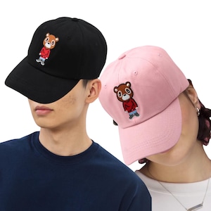 Op de afbeelding: Twee baseballpetten, één zwart en één roze, elk met een geborduurd berenontwerp. De beer draagt een rode jas en een blauwe spijkerbroek. De petten hebben een gebogen klep en een verstelbare band.