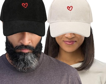 May include: Two corduroy baseball caps, one black and one off-white, each embroidered with a small red heart. The caps have a classic six-panel design with a curved brim, suitable for casual wear.