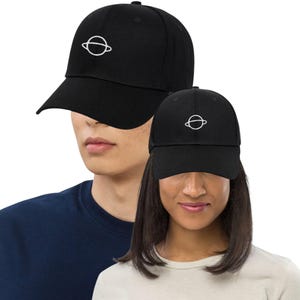 May include: Two black baseball caps, each featuring a white planet design. The caps are worn by two people. The caps have a curved peak and an adjustable strap at the back for a comfortable fit.
