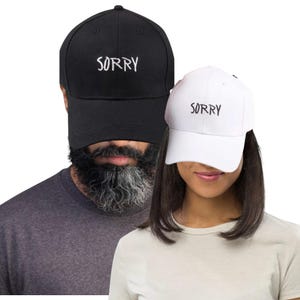 May include: Black and white baseball caps with the word "SORRY" embroidered on the front. The black cap is worn by a person with a beard, and the white cap is worn by a person with dark hair. The caps are a casual style.