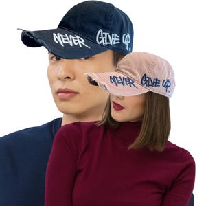 May include: Two baseball caps with the words "NEVER GIVE UP" in white script. One cap is navy blue, the other is a light pink. Both caps have a distressed, worn look. The caps are on a white background.